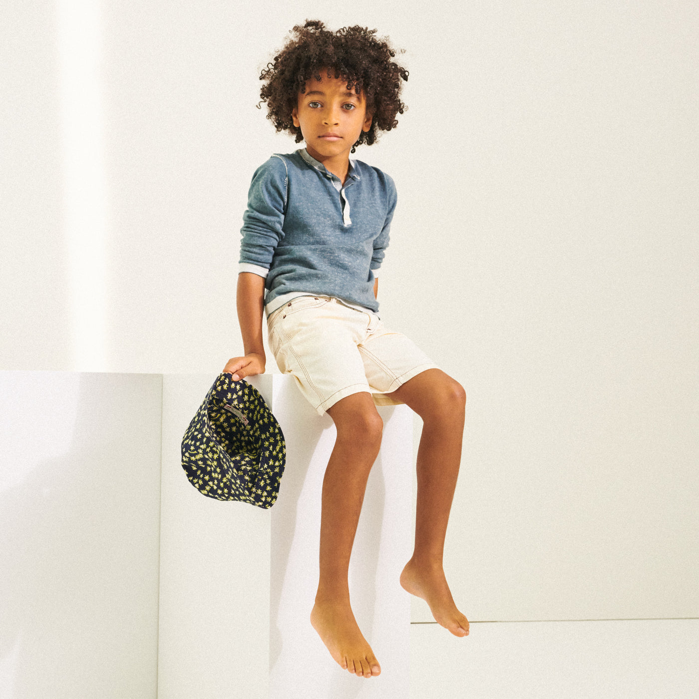 Boy sitting in blue shirt and holding a printed hat from Bonpoint Spring Summer 2022 Collection