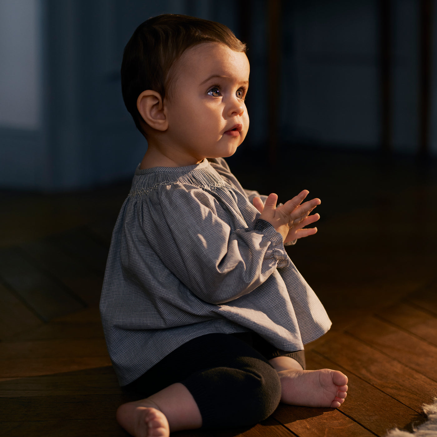 Bonpoint baby sitting on the hardwood floor wearing a blue blouse