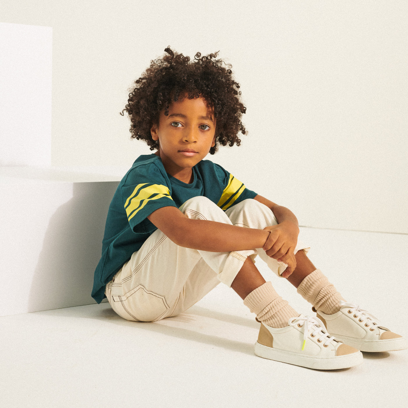 Boy sitting in green t-shirt and cream pants from Bonpoint Spring Summer 2022 Collection