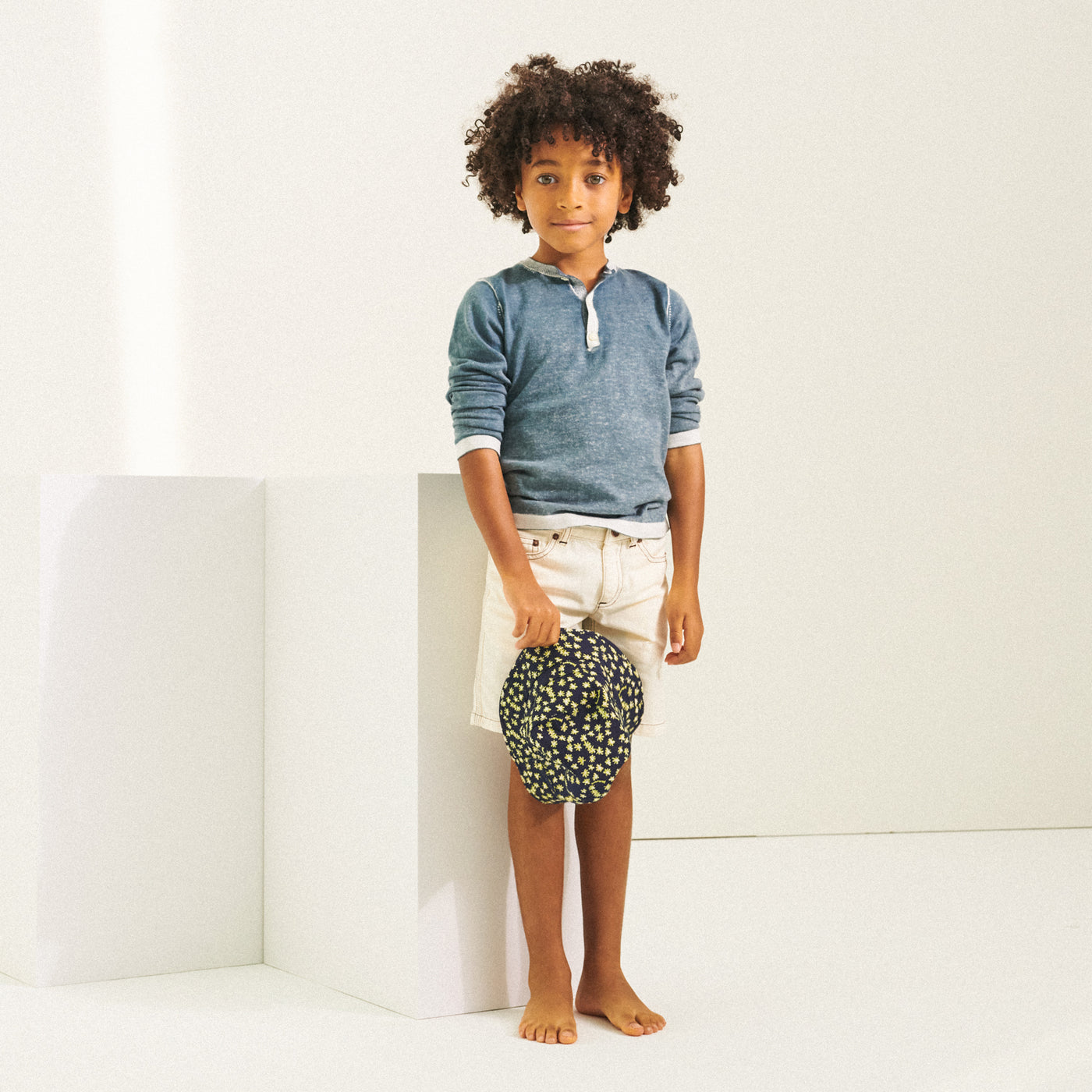 Boy standing in blue shirt and white shorts while holding a printed hat from Bonpoint Spring Summer 2022 Collection
