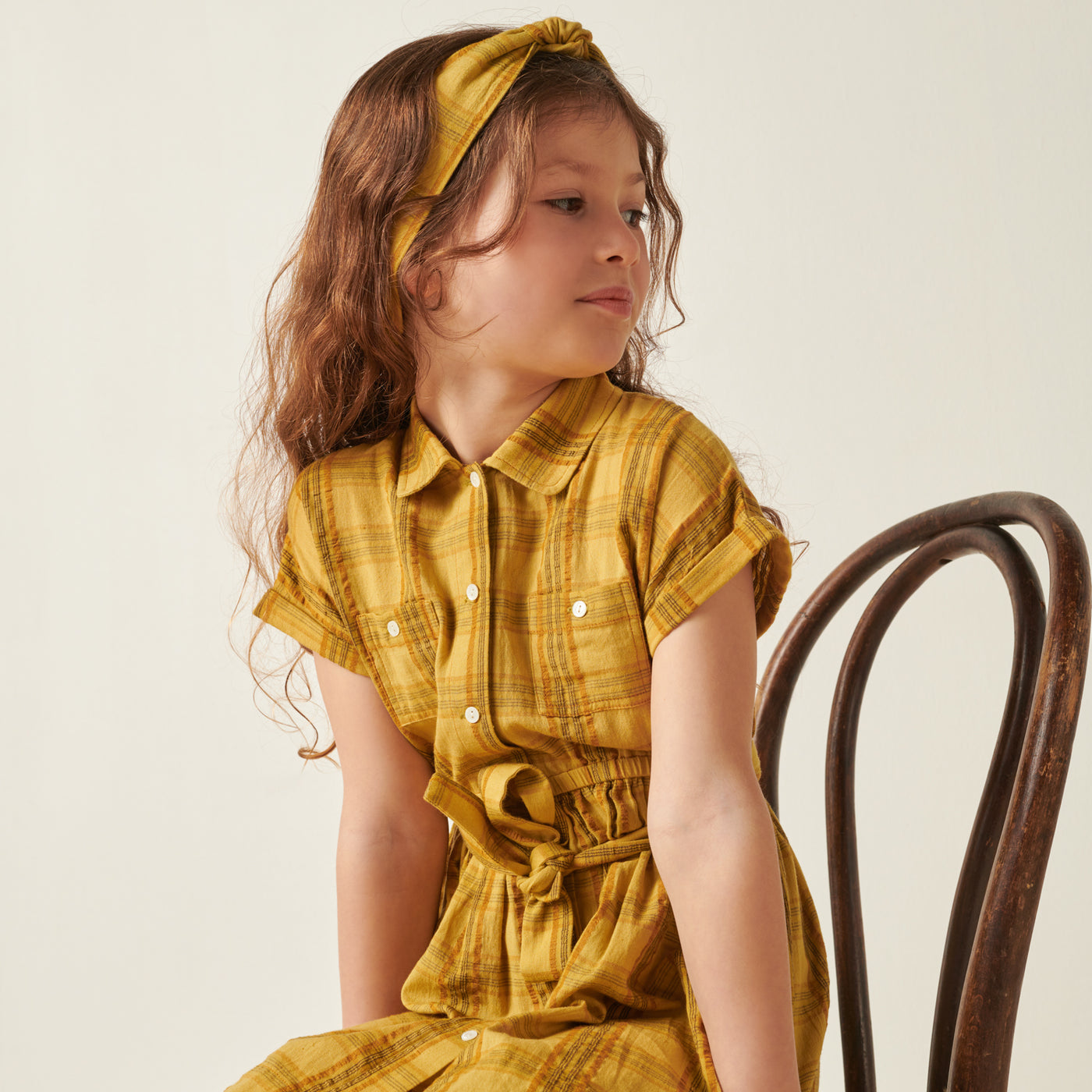 Girl sitting in chair wearing yellow dress with plaid design from Bonpoint Pre-Fall 2022 Collection
