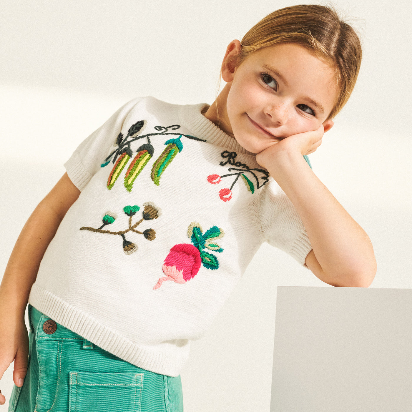 Girl leaning on table in brightly stitched graphic shirt from Bonpoint Spring Summer 2022 Collection