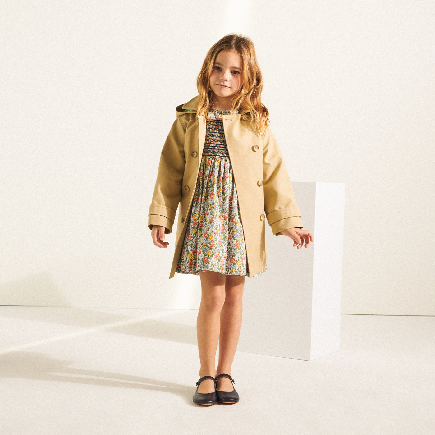 Girl standing in floral dress with beige trench coat from Bonpoint Spring Summer 2022 Collection