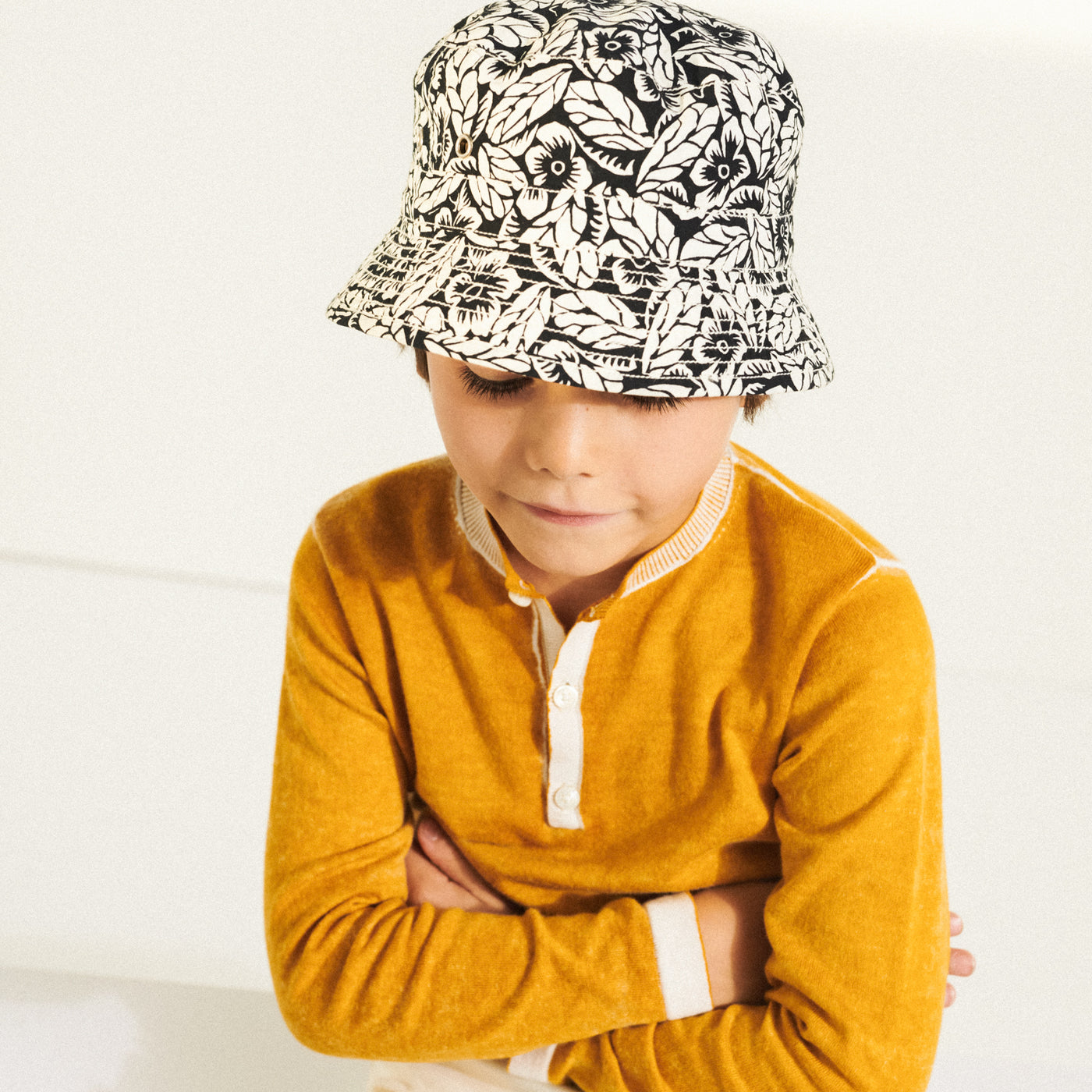 Boy with arms crossed wearing a yellow shirt and black and white printed hat from Bonpoint Spring Summer 2022 Collection